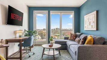 Ein modernes Wohnzimmer mit einem grauen Sofa, einem Couchtisch, einem Schreibtisch mit Stuhl, einem an der Wand montierten Fernseher und einem großen Fenster mit Stadtblick.