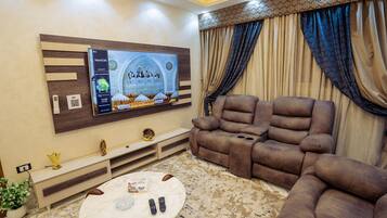 A living room with a flat-screen TV mounted on the wall, a brown sofa, a small coffee table, and a patterned rug.