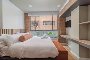 A modern bedroom with a large bed, a wooden headboard, a bench, a TV mounted on the wall, and a view of a brick building through a window. - Apto Moderno 1 hab Bog Sierra Madre (Bogotá)
