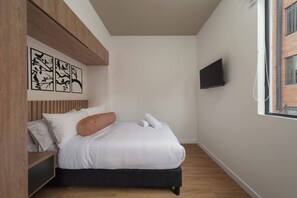 A bedroom with a bed, a wooden headboard, a flat-screen TV mounted on the wall, and a window with a view of a brick building. - Studio Bogota 1Bed Sierra Madre 306 (Bogotá)