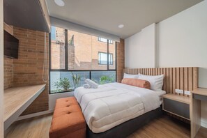 A modern bedroom with a large window, a bed with white bedding, a wooden bench, and a desk. - Apto 1 Hab Ed sierra Madre 209 (Bogotá)