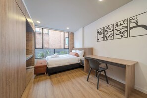 Ein modernes Schlafzimmer mit Bett, Schreibtisch und Stuhl, einem Fenster mit Blick auf ein Backsteingebäude und Wandbildern.