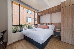 A modern bedroom with a large window, a bed with white bedding, a wooden headboard, and a built-in wardrobe. - Studio Bogota 1Bed Sierra Madre 305 (Bogotá)