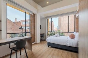 Un dormitorio con cama, escritorio y silla. Amplias ventanas ofrecen vistas de edificios.