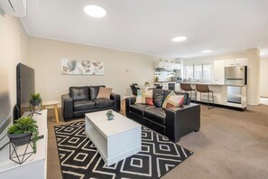 Una sala de estar moderna con sofá, mesa de centro y televisor. Hay una zona de cocina con refrigerador y mesa de comedor.