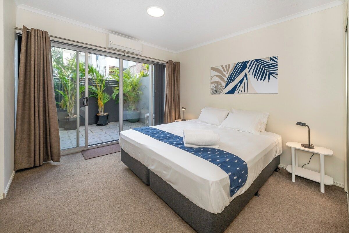 A bedroom with a bed, a bedside table, a window with curtains, and a view of the outside.