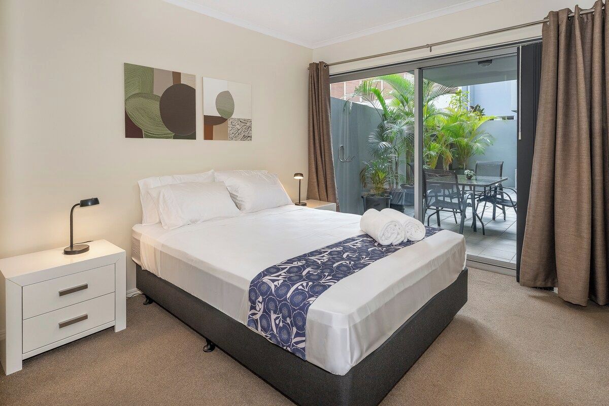 A hotel room with a bed, a nightstand, a lamp, a sliding glass door to an outdoor area with a table and chairs, and artwork on the wall.