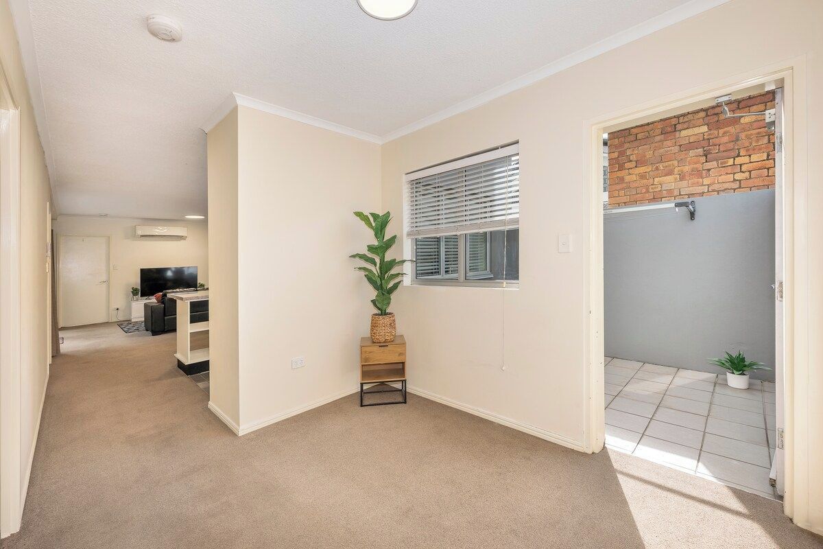 A room with a carpeted floor, a small wooden table with a plant, a window with blinds, and a door leading to a balcony with a tiled floor.