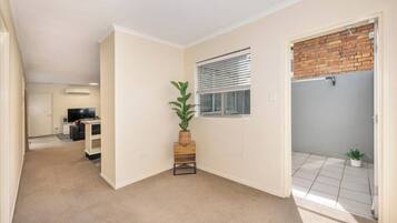 A room with a carpeted floor, a small wooden table with a plant, a window with blinds, and a door leading to a balcony with a tiled floor.