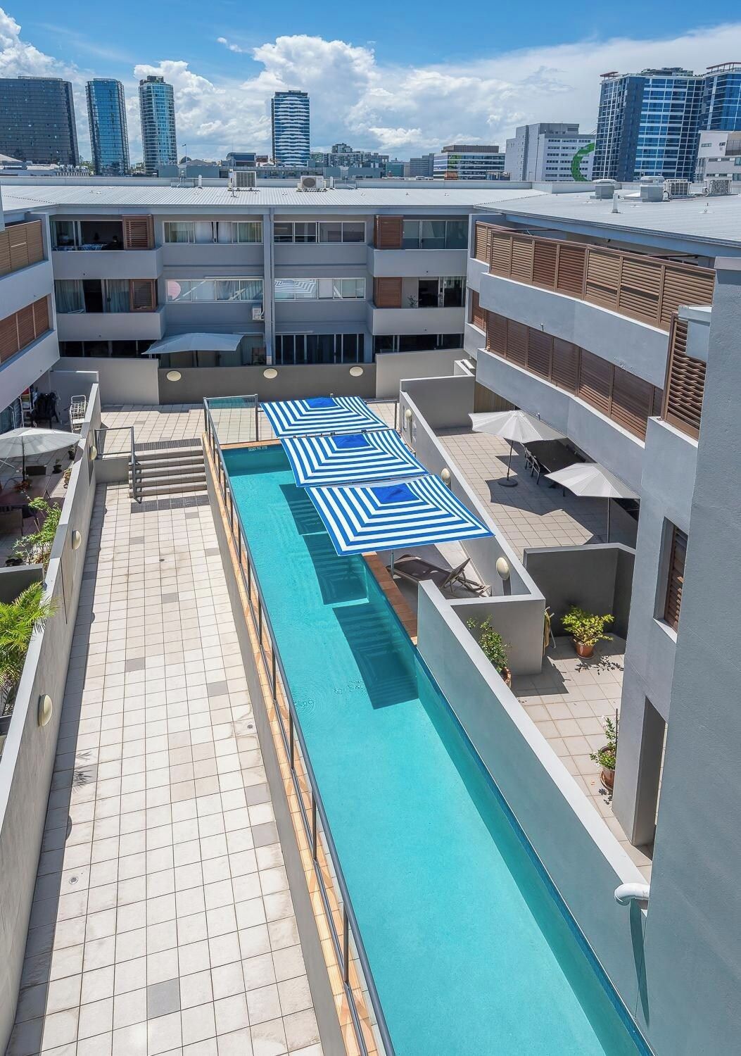 A rooftop swimming pool with blue and white striped umbrellas, surrounded by modern buildings.