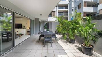 A modern outdoor dining area with a table and chairs, surrounded by potted plants and a view of a residential building.