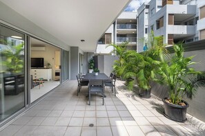 Uma área moderna para refeições ao ar livre, com mesa e cadeiras, cercada por plantas em vasos e vista para um prédio residencial.