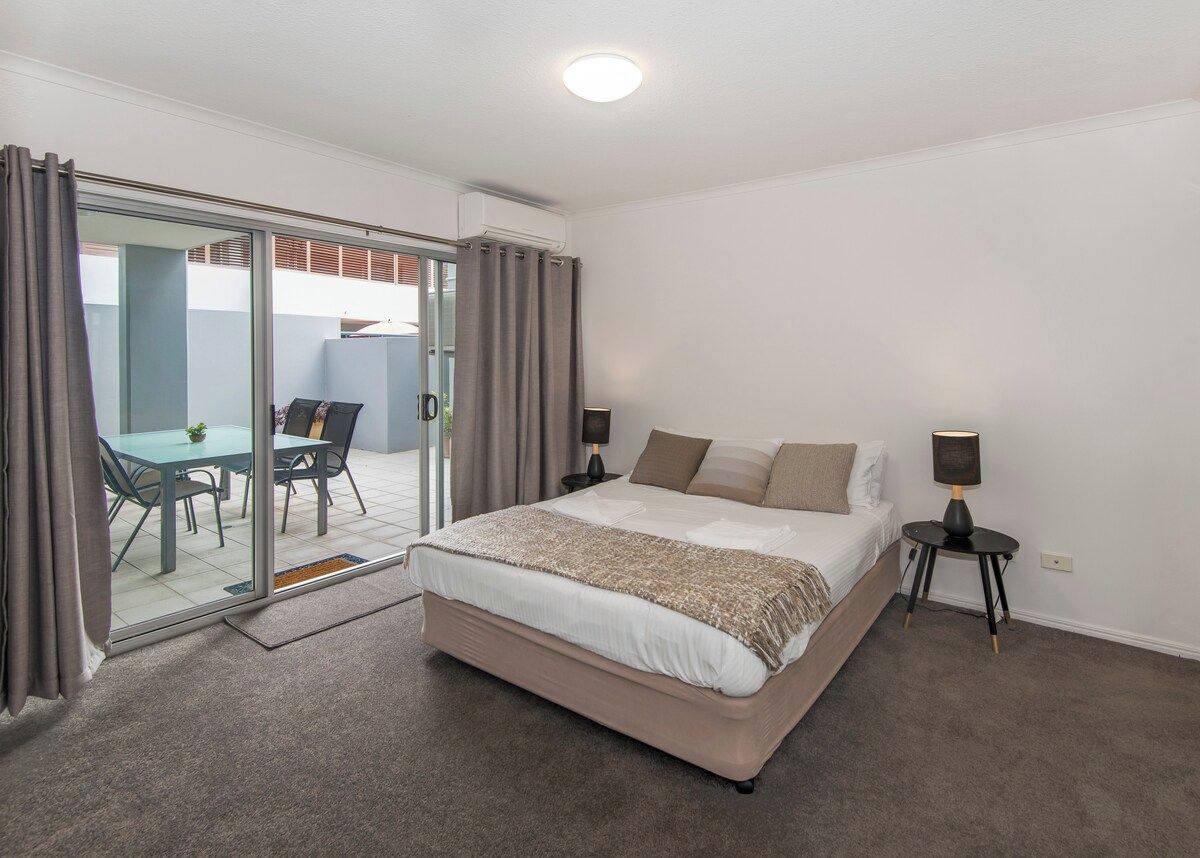 A bedroom with a large bed, a bedside table, a lamp, and a sliding glass door leading to a balcony with a table and chairs.