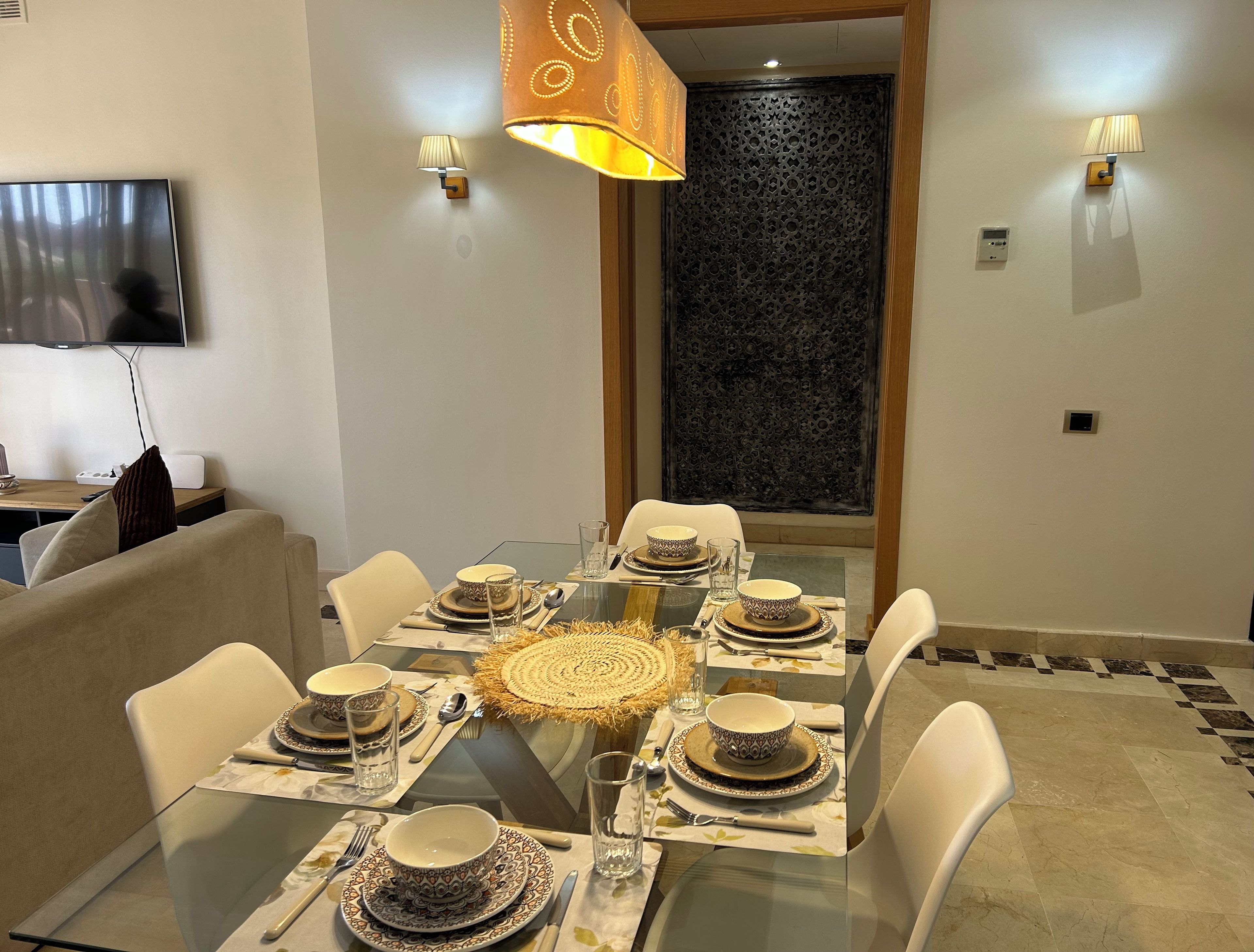 Un comedor con una mesa de vidrio preparada para seis personas, rodeada de sillas blancas, con un televisor montado en la pared a la izquierda.