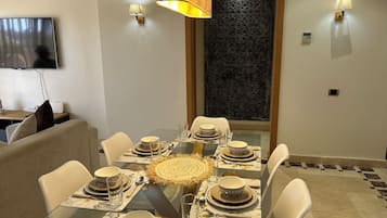 Un coin repas avec une table en verre dressée pour six personnes, entourée de chaises blanches, et un téléviseur fixé au mur sur le côté gauche.