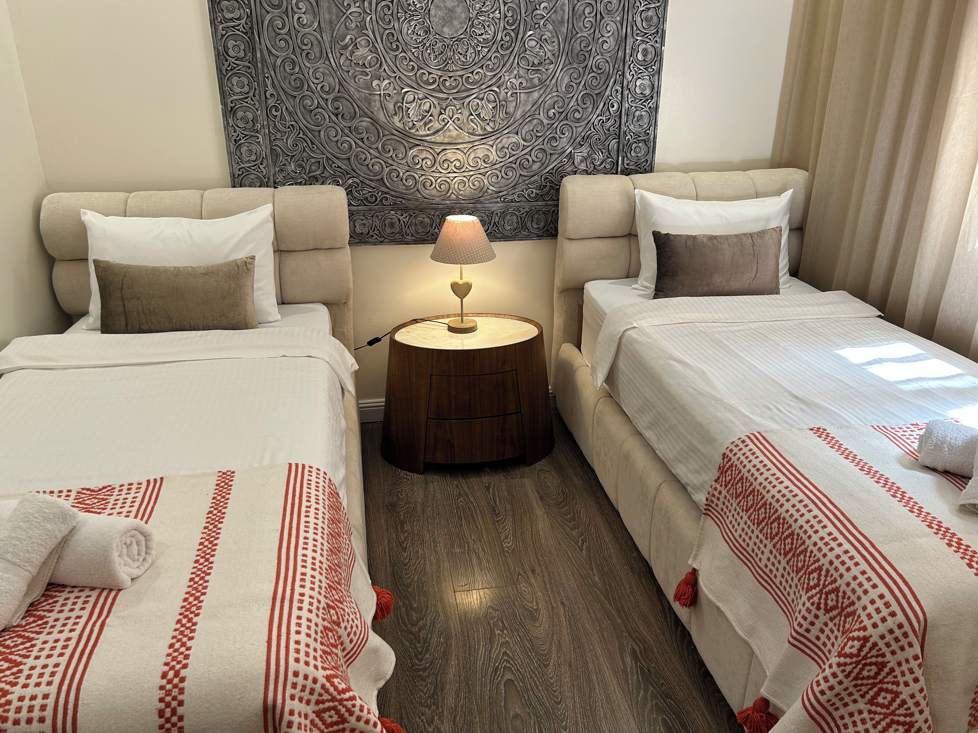 A hotel room with two beds, a round wooden nightstand with a lamp, a tapestry on the wall, and a window with curtains.