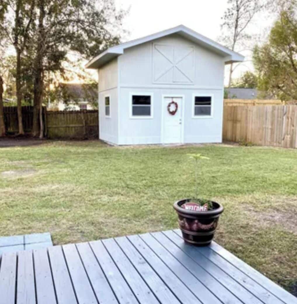 A small, single-story building with a white exterior and a garage door. There is a wooden deck in the foreground with a planter on it.