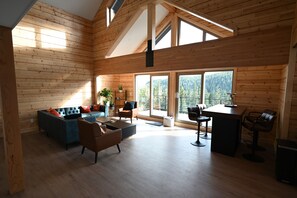 A spacious room with a high ceiling, wooden walls, and large windows offering a view of a forested area. - Sabre Tooth Lodge - Luxury Stay on Mt. Washington. Hot Tub, Sauna, on site (Mt. Washington, Comox Valley)