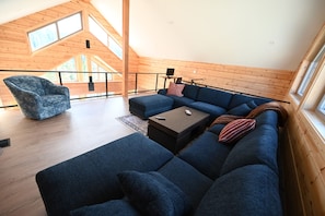 A modern loft-style room with a high ceiling, wooden beams, a large sectional sofa, a single armchair, and a small table. - Sabre Tooth Lodge - Luxury Stay on Mt. Washington. Hot Tub, Sauna, on site (Mt. Washington, Comox Valley)