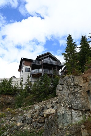 Un bâtiment moderne à plusieurs étages, doté d’un balcon imposant, entouré de végétation naturelle et d’un terrain rocheux.