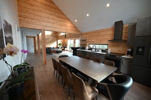 A modern kitchen with wooden paneling, a dining area with a long table and chairs, and a view of the outdoors. - Sabre Tooth Lodge - Luxury Stay on Mt. Washington. Hot Tub, Sauna, on site (Mt. Washington, Comox Valley)