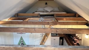 Quarto em estilo loft com teto inclinado, vigas de madeira, uma cama e um pequeno espaço para escritório.