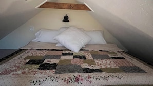 Quarto com teto inclinado, vigas de madeira, cama com colcha e abajur.