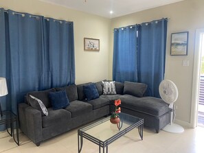 A living room with a sectional sofa, a glass coffee table, a fan, and blue curtains. - Modern 1BR Apt at The Aqueducts - Gated, Pool, Walk to Beach, Centrally Located (Westmoreland)