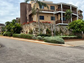A multi-story building with a stone facade, balconies, and a landscaped garden with trimmed bushes and palm trees. - Modern 1BR Apt at The Aqueducts - Gated, Pool, Walk to Beach, Centrally Located (Westmoreland)