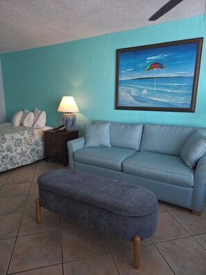 A room with a teal wall, a painting of a beach scene, a sofa, a bench, and a bedside table with a lamp. - Oceanfront Paradise at Fountain Beach Resort in gorgeous Daytona Beach (Daytona Beach Shores)