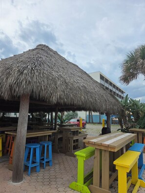 Un área de comedor frente a la playa con sombrillas de paja, mesas de madera y taburetes de colores.