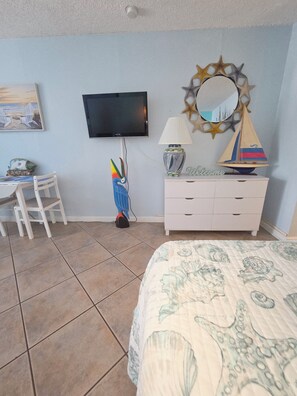 Un dormitorio con cama, escritorio, silla, televisor, lámpara y un espejo decorativo.