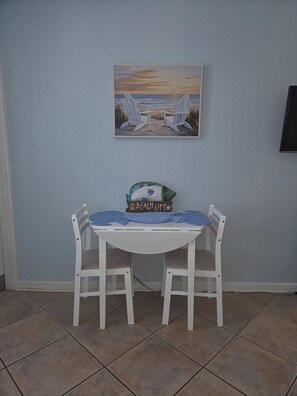 Un comedor con una mesa y sillas blancas, un cuadro con temática marina arriba y una pequeña señal en la mesa.