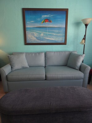 Una habitación con pared azul claro, un cuadro enmarcado de una escena de playa, un sofá gris con cojines y un reposapiés gris.
