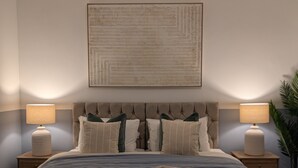 Une chambre à coucher avec un grand lit, deux lampes de chevet, une tête de lit capitonnée et un tableau abstrait accroché au mur.