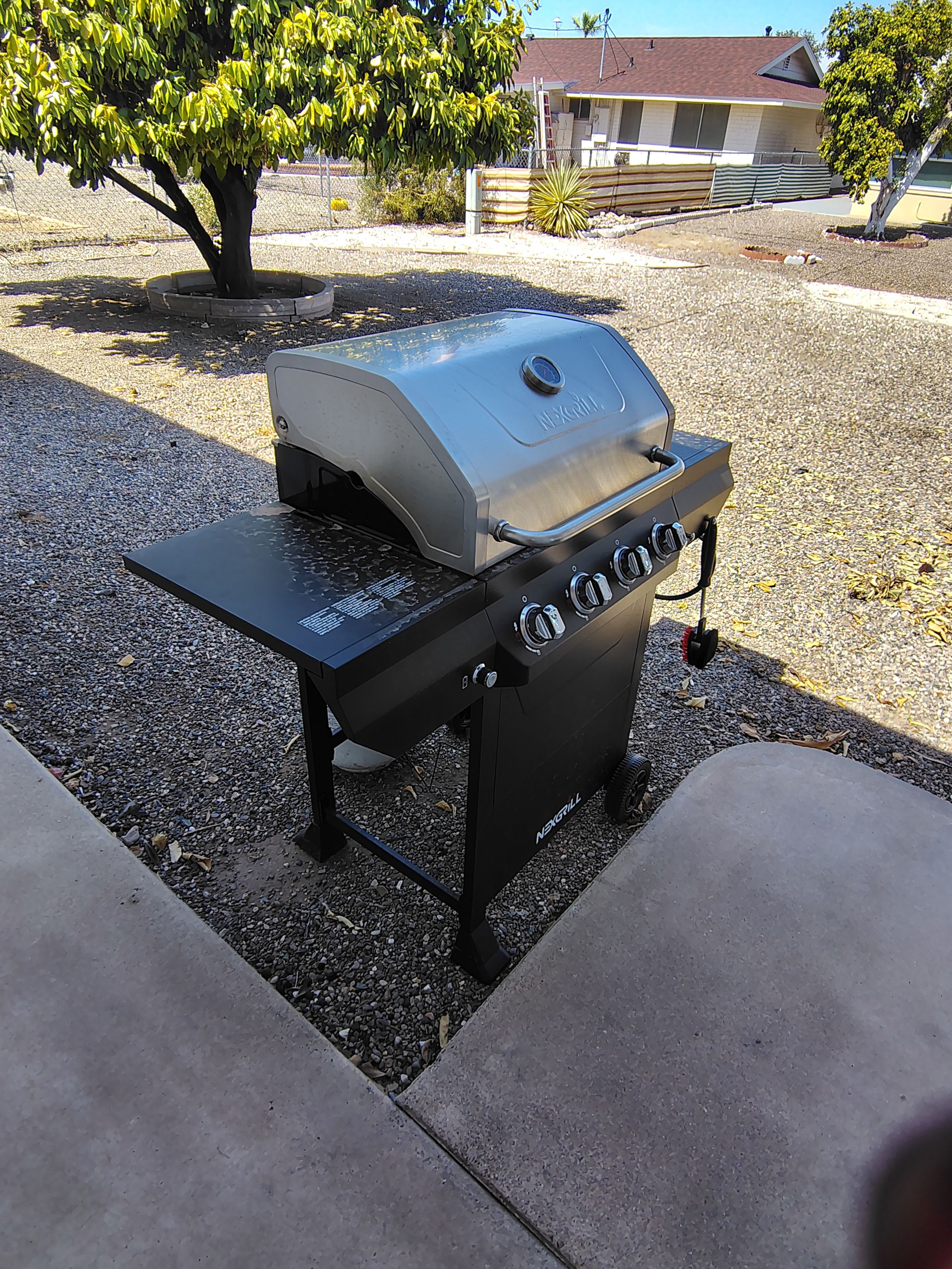 A black grill with four burners and a side shelf, placed on a concrete patio with gravel around.