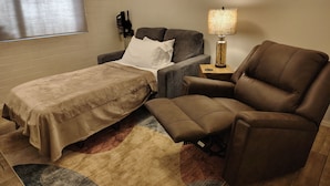 Quarto de hotel com cama, sofá, poltrona, luminária e janela com persianas.