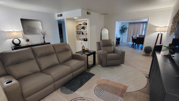 Uma sala de estar moderna com sofá, poltrona e mesa de centro, uma estante e um espaço de jantar ao fundo.