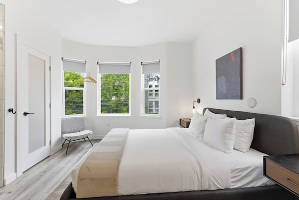 A modern bedroom with a large bed, a bedside table, a nightstand, and a window with a view of greenery.