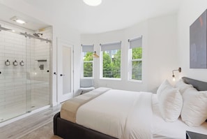 A modern bedroom with a glass-enclosed shower, a bed with white bedding, and two windows offering a view of greenery.