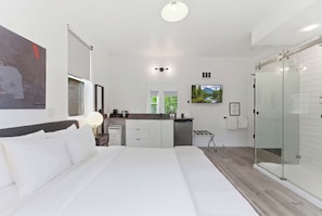 A modern hotel room with a large bed, a kitchenette, a glass shower enclosure, and a view of greenery.