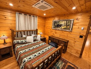 Ein Schlafzimmer einer Holzhütte mit einem Bett, Nachttischen, einer Lampe, einem Gemälde und einem Fenster mit Vorhängen.