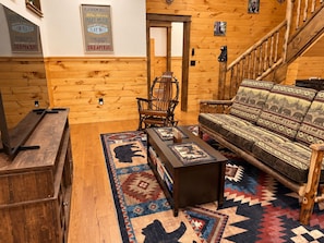 Ein gemütliches Zimmer mit Holzboden, einer Couch mit gemusterten Kissen, einem kleinen Tisch mit dekorativer Teppich und einer Treppe mit Holzwand.