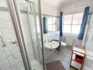 Un baño con ducha envuelta en vidrio, lavamanos, inodoro y ventana con cortinas.