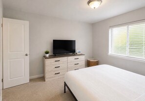 A bedroom with a bed, a television on a dresser, a window with blinds, and a door. - Peaceful 3-bedroom house in charming Yelm perfect for relaxing getaway (Yelm)