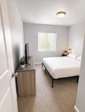 A bedroom with a bed, a desk with a television, a window with blinds, and a ceiling light. - Peaceful 3-bedroom house in charming Yelm perfect for relaxing getaway (Yelm)