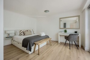 Un dormitorio con cama, mesita de noche, lámpara, planta en la mesita, escritorio, silla y espejo.