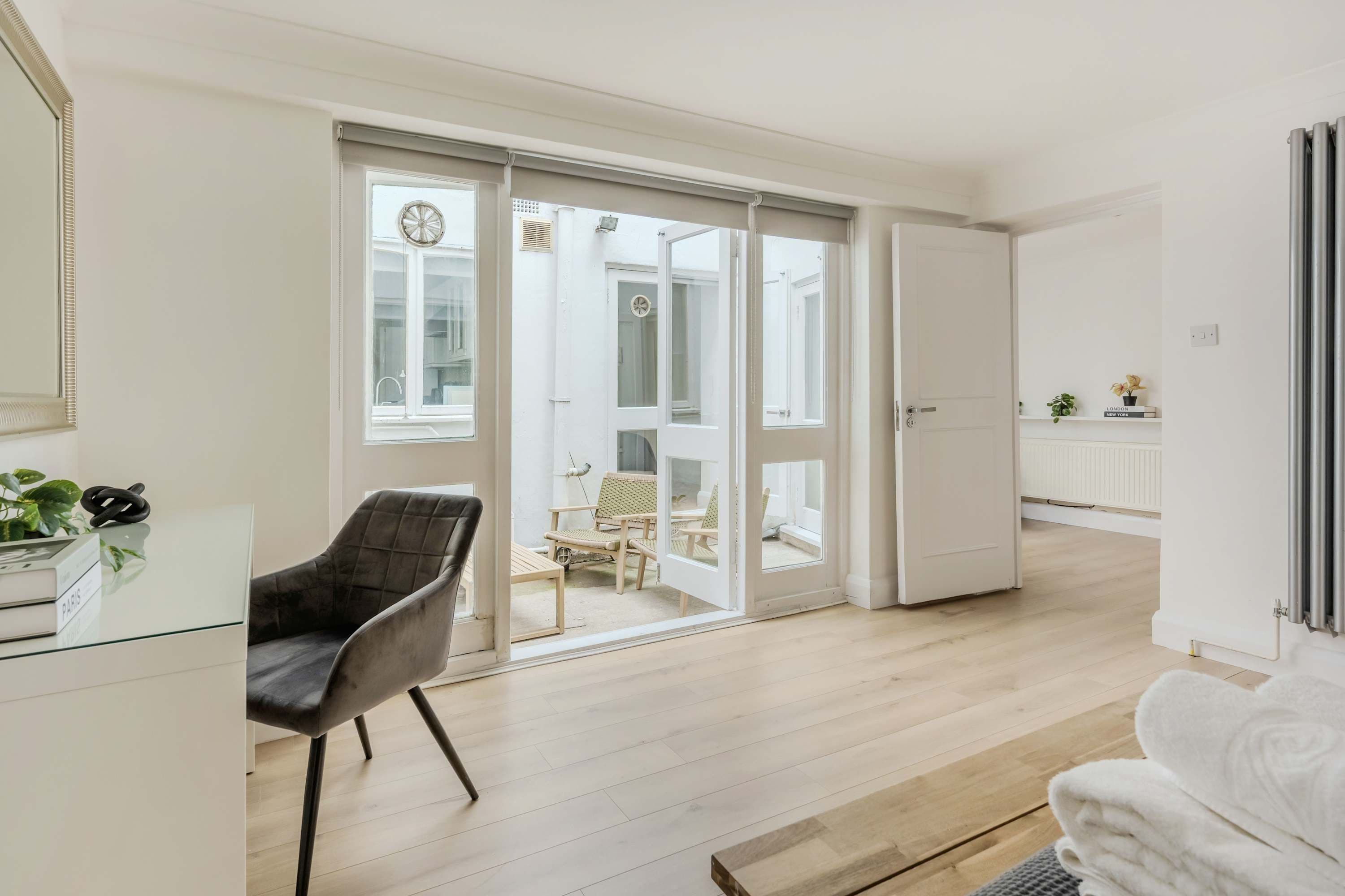 A modern living room with a glass desk, a black chair, and a white sofa. There is a sliding glass door leading to a balcony with outdoor furniture.