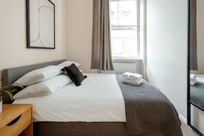 Ein Hotelzimmer mit Bett, Nachttisch, einem Fenster mit Vorhängen und Blick auf Gebäude.