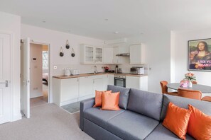 A modern kitchen with white cabinets, a grey sofa with orange cushions, and a dining area with a table and chairs. - The Foley Residence - Fitzrovia - by Frankie (London)
